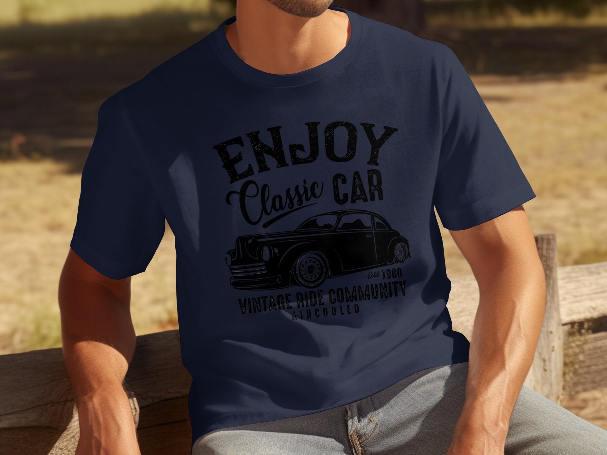 A person sits on a wooden bench wearing a navy blue t-shirt by Garment Graphics, featuring a vintage car graphic with the text Enjoy Classic Car, Vintage Ride Community, Aircooled, Est. 1980, ideal for car enthusiasts who love timeless journeys.