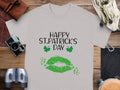 A Garment Graphics gray graphic T-shirt lies on a wooden surface, surrounded by a camera, leather items, binoculars, and headphones. It features vibrant green lips and clovers with the festive message Happy St. Patricks Day.