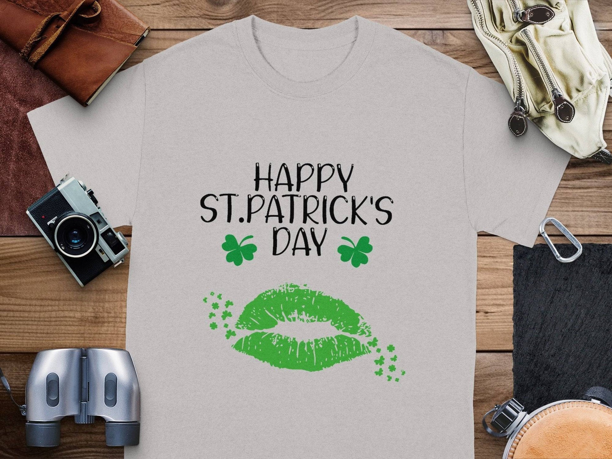 A Garment Graphics gray graphic T-shirt lies on a wooden surface, surrounded by a camera, leather items, binoculars, and headphones. It features vibrant green lips and clovers with the festive message Happy St. Patricks Day.