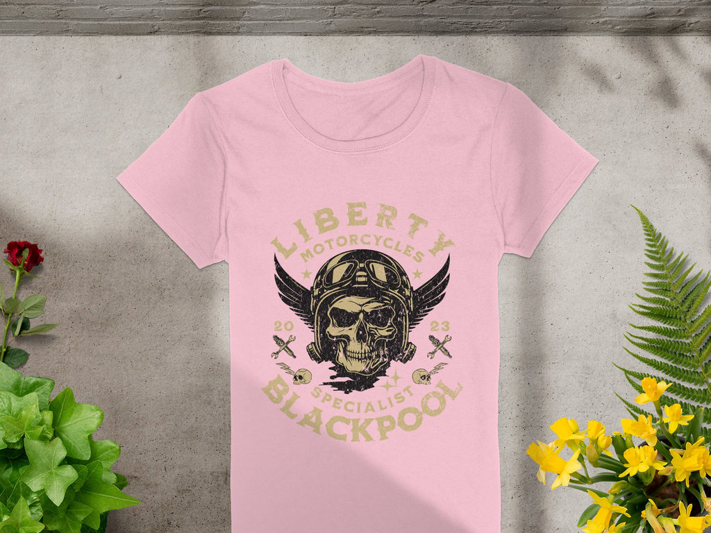The Garment Graphics Blackpool-themed T-shirt features a Liberty Motorcycles Specialist Blackpool 2023 design with a striking skull and wings motif. Displayed on concrete, its beautifully framed by lush green plants and vibrant yellow flowers.