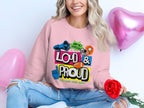 Wearing a pink Loud & Proud sweatshirt by Garment Graphics, known for its unique style and unisex sizing, someone sits with a red rose among pink heart-shaped balloons. Theyre smiling in ripped jeans, exuding a confident and joyful aura.