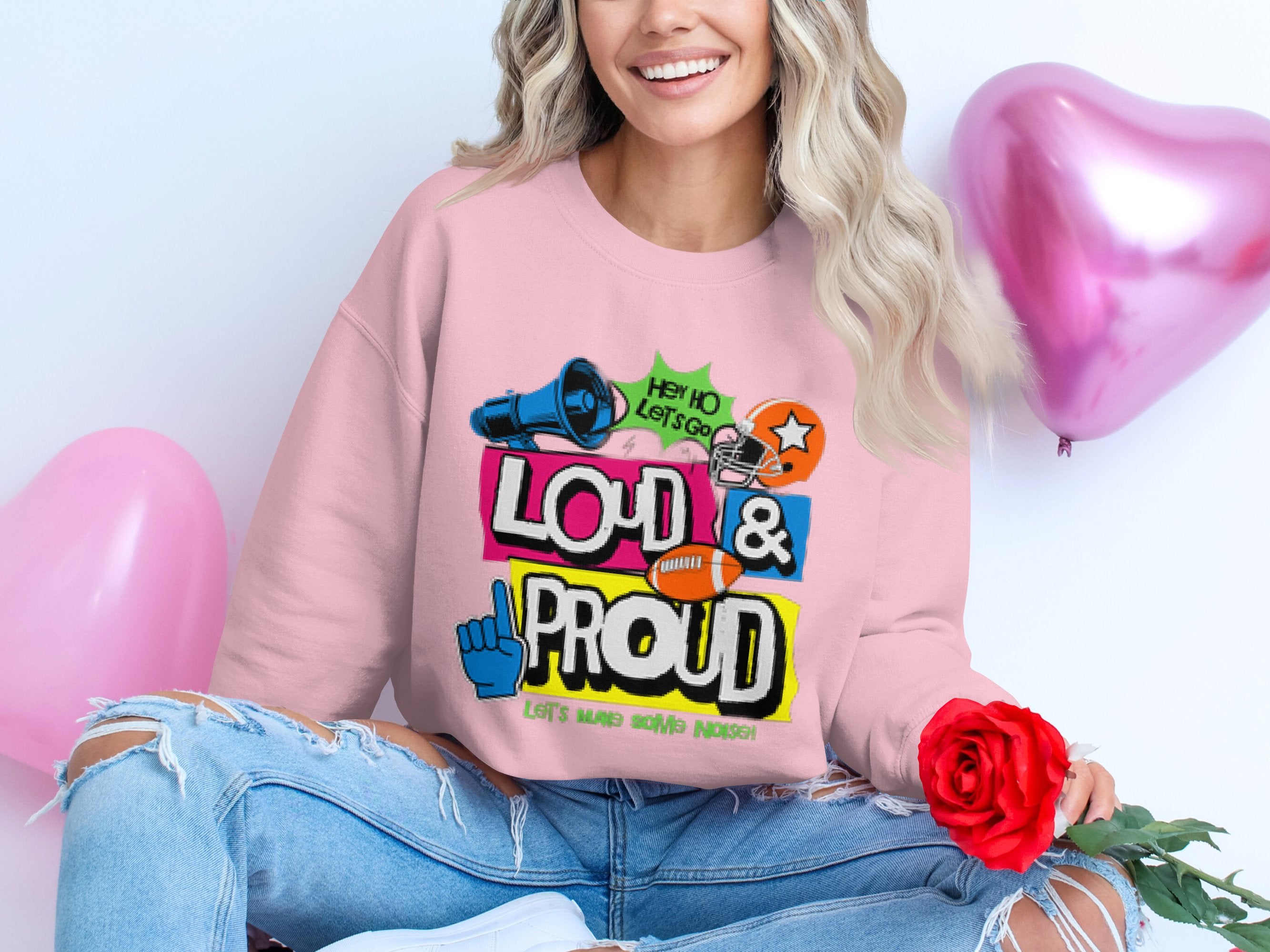 Wearing a pink Loud & Proud sweatshirt by Garment Graphics, known for its unique style and unisex sizing, someone sits with a red rose among pink heart-shaped balloons. Theyre smiling in ripped jeans, exuding a confident and joyful aura.