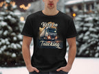 In a snowy setting, a man showcases Garment Graphics black T-shirt designed for truck enthusiasts. The graphic features a large truck on a road with the phrase Keep on Trucking, against a backdrop of snow-covered trees and twinkling lights.