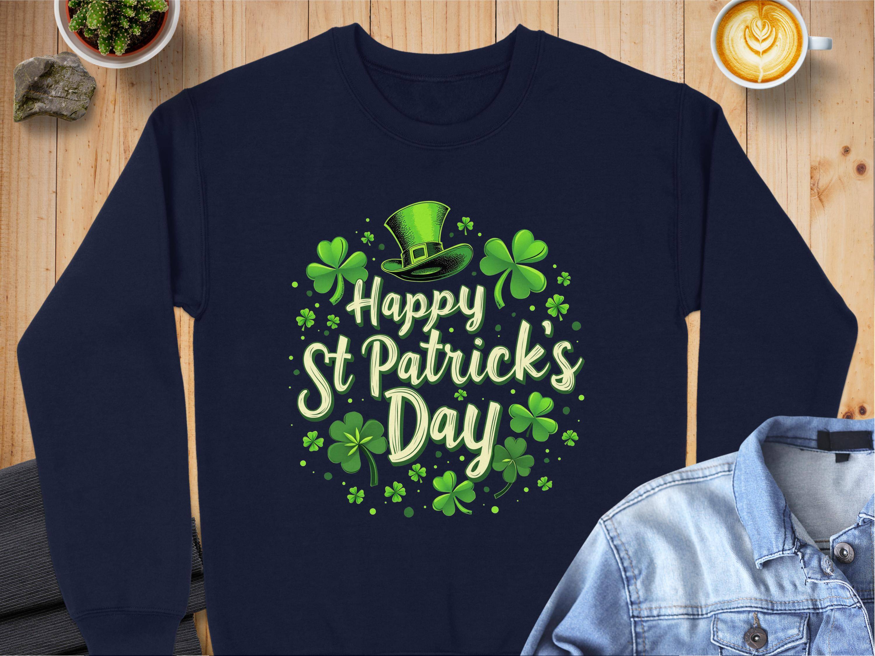 A Garment Graphics navy sweatshirt with a shamrock, Happy St. Patricks Day design, green clovers, and a leprechaun hat sits on wooden surface by a denim jacket and latte, adding festive cheer to your celebration.