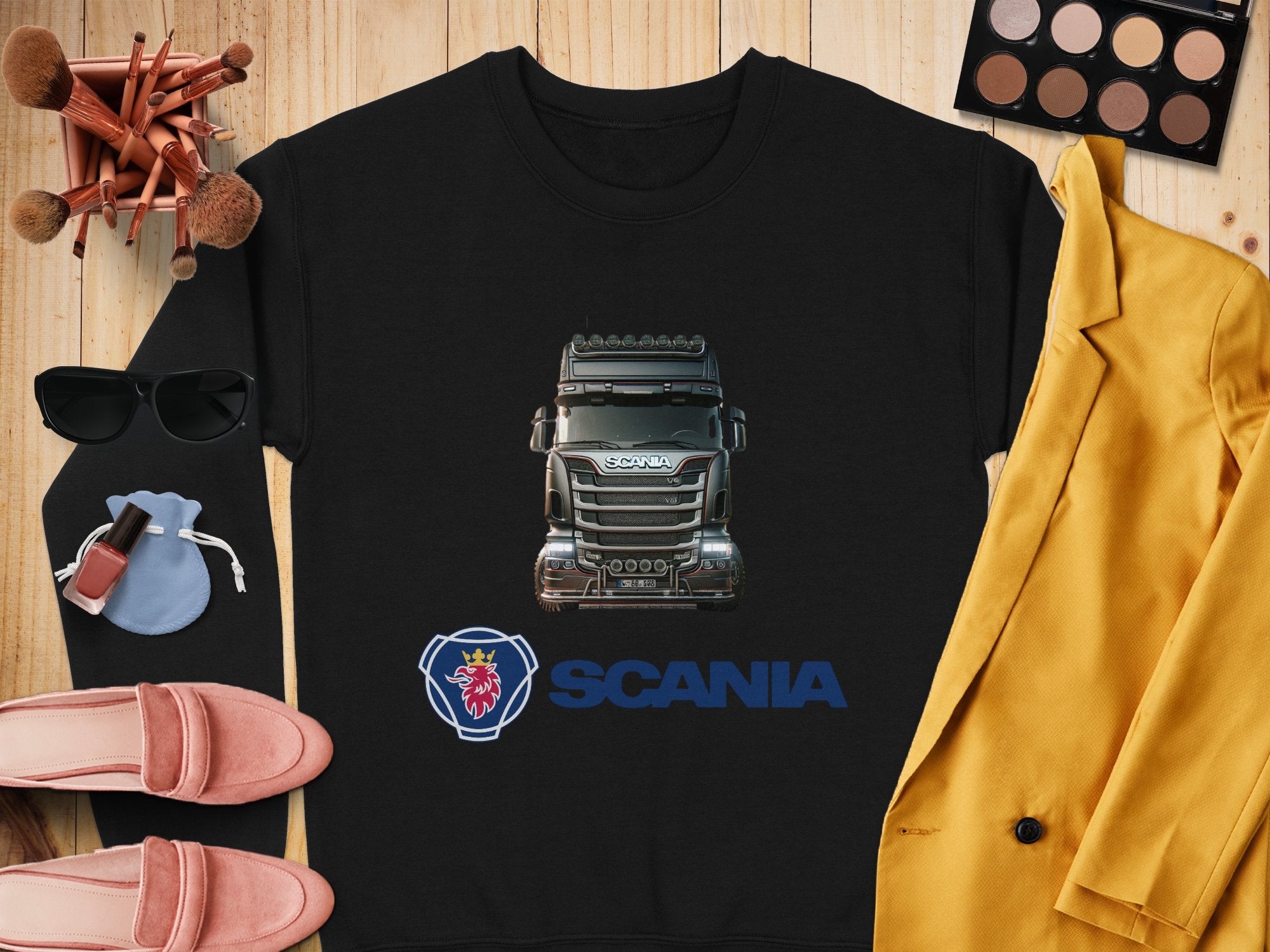 Laid on a wooden surface is a black crewneck sweatshirt featuring a Scania truck design by Garment Graphics. Perfect for truck lovers, its paired with items like a yellow blazer, pink loafers, sunglasses, makeup brushes, an open eyeshadow palette, and a lipstick-topped compact mirror.