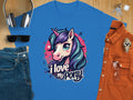 The Garment Graphics T-shirt, adorned with a vibrant blue cartoon unicorn and the text I love my pony, is displayed on a wooden surface alongside headphones, keys, sunglasses, blue jeans, an orange button-up shirt, and a wristwatch.