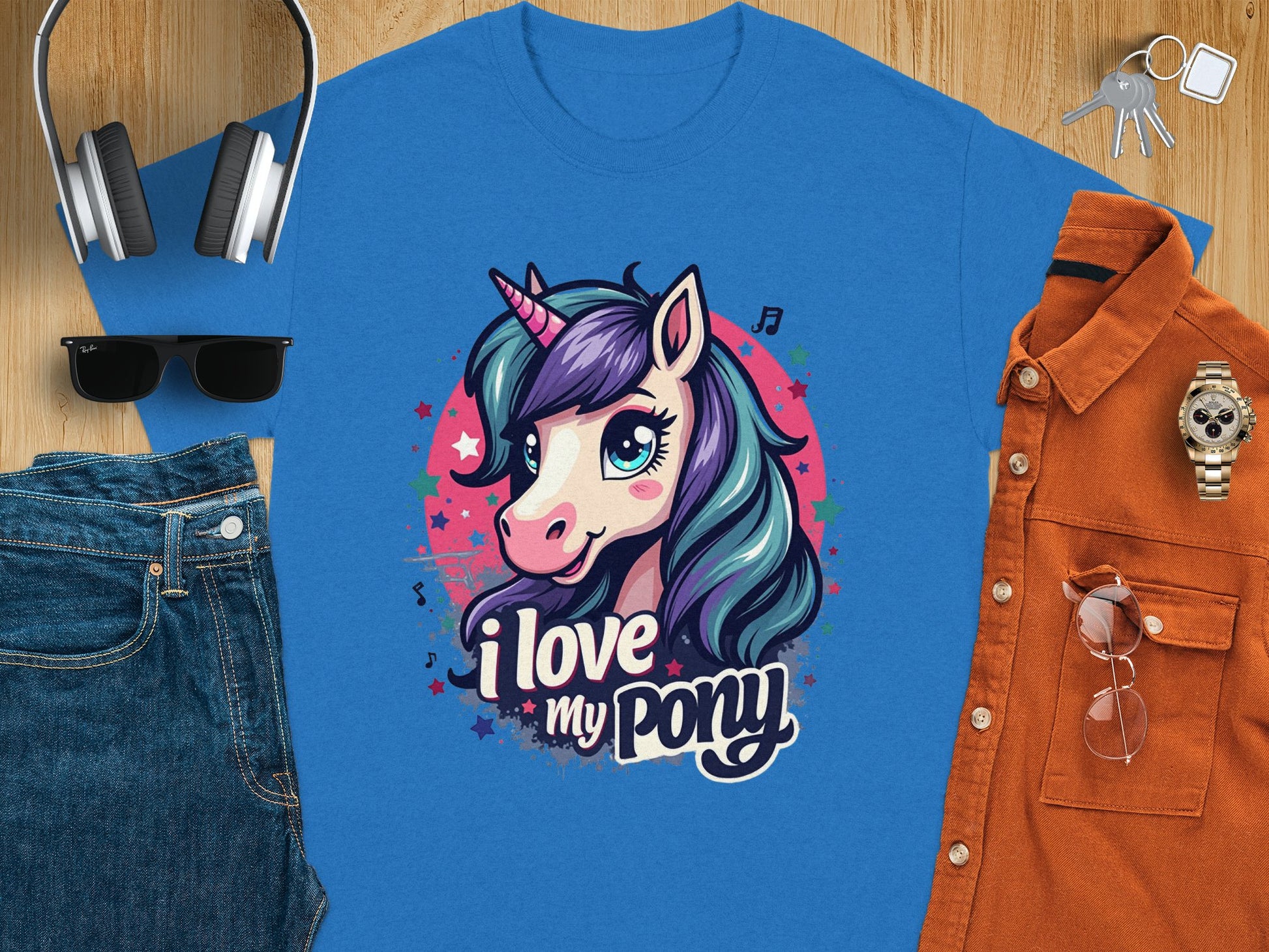 The Garment Graphics T-shirt, adorned with a vibrant blue cartoon unicorn and the text I love my pony, is displayed on a wooden surface alongside headphones, keys, sunglasses, blue jeans, an orange button-up shirt, and a wristwatch.