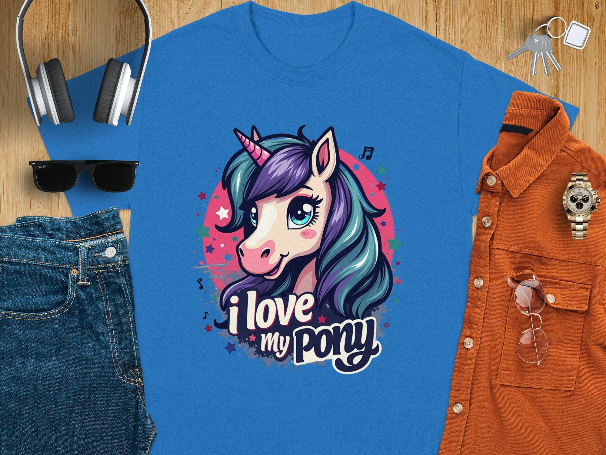 The Garment Graphics T-shirt, adorned with a vibrant blue cartoon unicorn and the text I love my pony, is displayed on a wooden surface alongside headphones, keys, sunglasses, blue jeans, an orange button-up shirt, and a wristwatch.