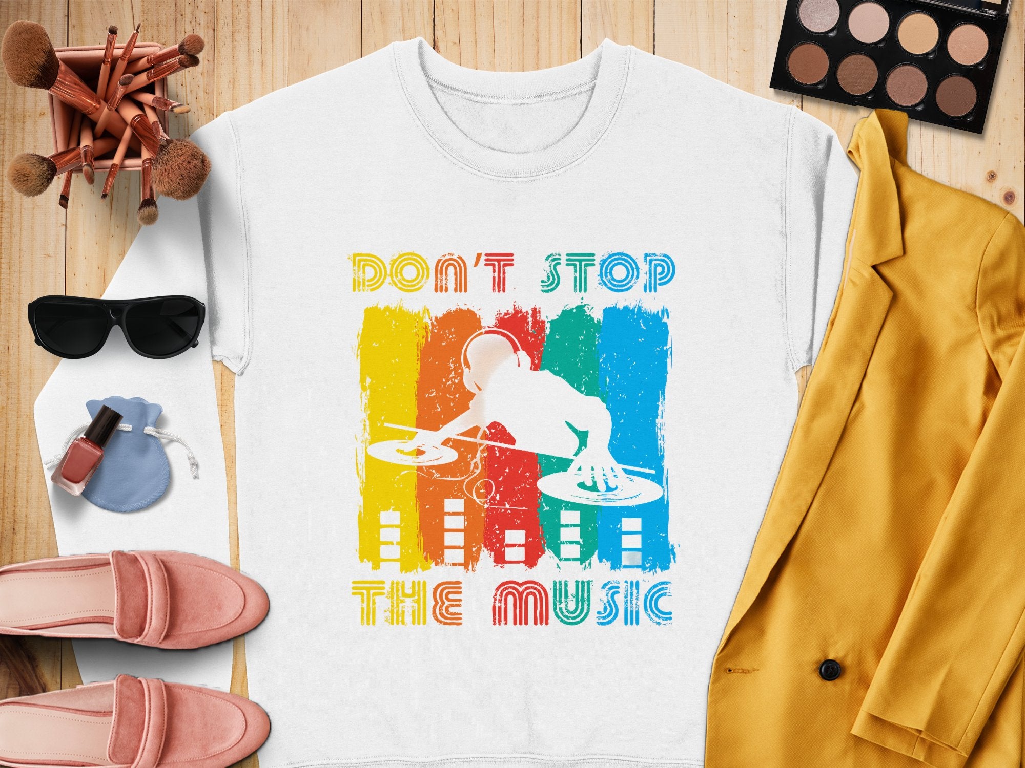 Garment Graphics offers a white sweatshirt with a vibrant DJ design and Dont Stop the Music phrase. Music lovers can pair it with a yellow jacket, pink shoes, sunglasses, makeup palette and brushes, nail polish, and earrings—all displayed on a wooden surface.