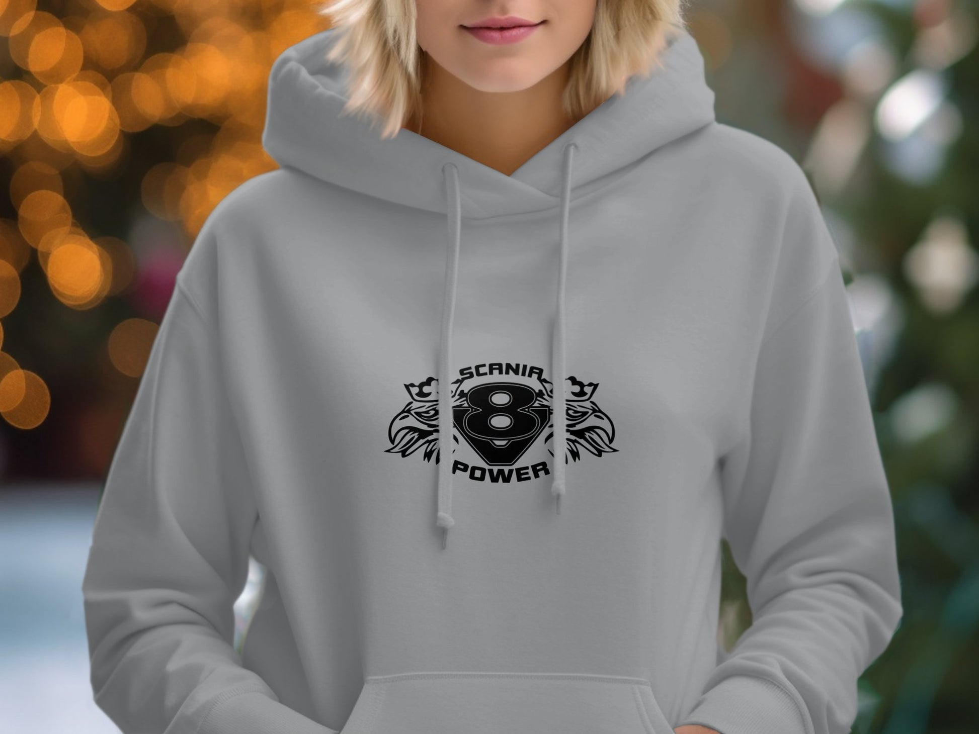 A person sports a Garment Graphics light gray hoodie with a bold SCANIA POWER emblem in classic fit, featuring a stylized 8 and decorative motifs, set against a background of subtly blurred warm bokeh lights.