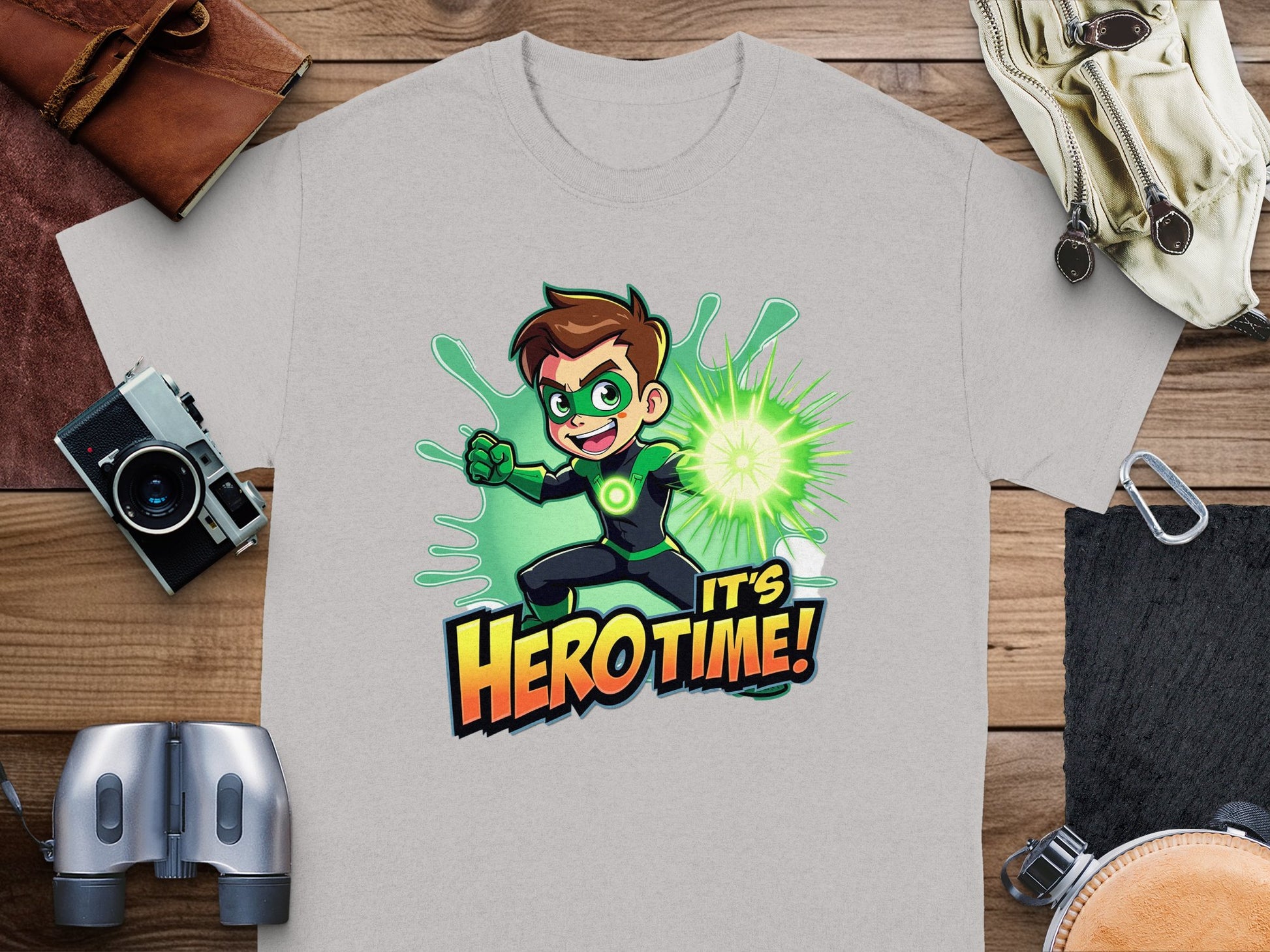 A light gray t-shirt by Garment Graphics features a cartoon character with brown hair in a green suit emitting glowing green light, with the text Its Hero Time! below. Ideal for superhero graphic tee fans, its displayed on a wooden surface amidst travel items.