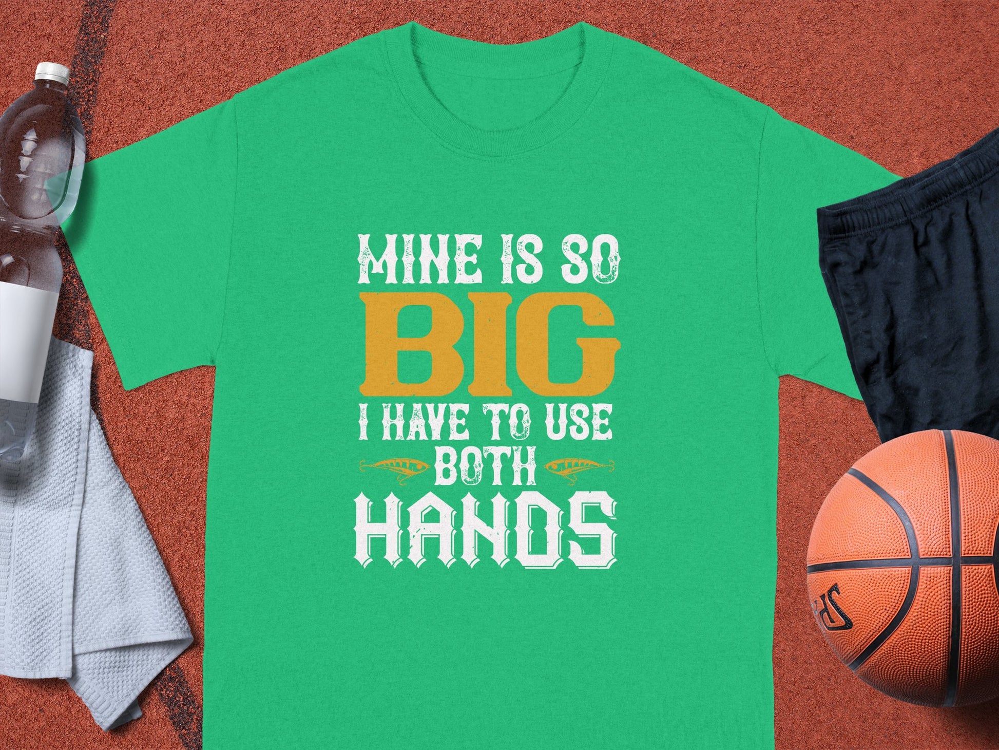 Garment Graphics green T-shirt boasts a funny design, MINE IS SO BIG I HAVE TO USE BOTH HANDS, in white and gold. The scene includes a basketball, black shorts, water bottle, and gray towel on an orange sports surface, making it a classic graphic tee for sports lovers.