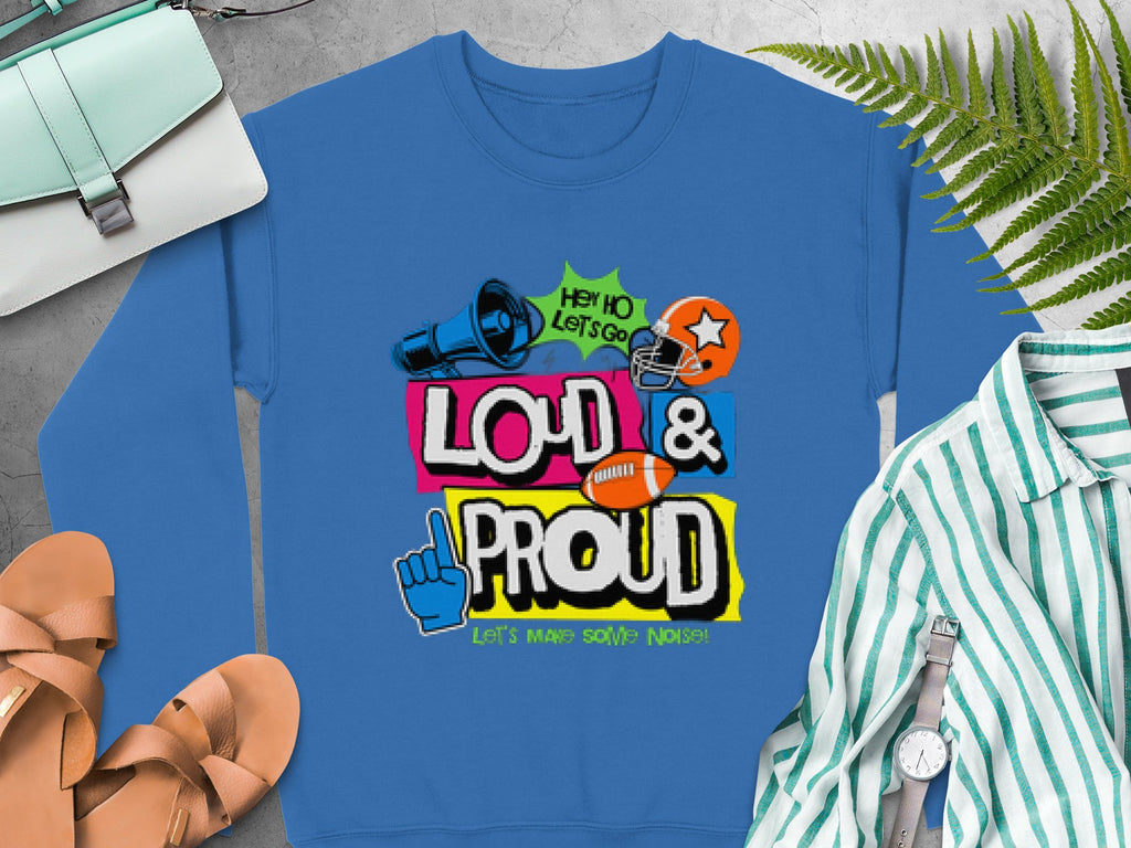 A blue sweatshirt from Garment Graphics lies on a surface next to sandals, a striped shirt, a green plant, and a purse. It features colorful text and graphics like a megaphone, football helmet, and foam finger. Unisex sizing is available.