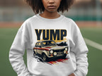 A person wears a white Garment Graphics sweatshirt featuring a YUMP design with a vintage rally car graphic. The blurred outdoor background of green and brown tones adds depth to the scene.