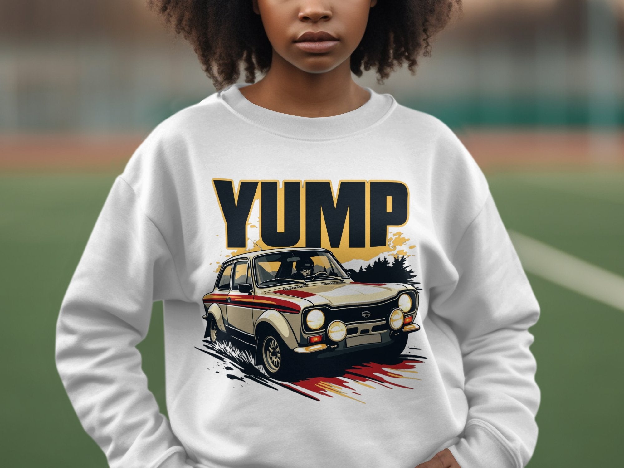 A person wears a white Garment Graphics sweatshirt featuring a YUMP design with a vintage rally car graphic. The blurred outdoor background of green and brown tones adds depth to the scene.