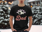 In a snowy outdoor setting, a person wears a Garment Graphics black graphic T-shirt with Soccer Dad and a soccer ball design. Snow-covered trees and softly lit lights complete the charming winter scene.