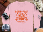 The unisex pink sweatshirt by Garment Graphics showcases orange BORN TO PLAY text above a soccer ball, with phrases like ONE GOAL - ONE DREAM, YOUNG SQUAD, FOOTBALL, and KICK, SCORE, CELEBRATE! Ideal for young squads, it is styled among a camera, leather bag, hat, binoculars, and rope.