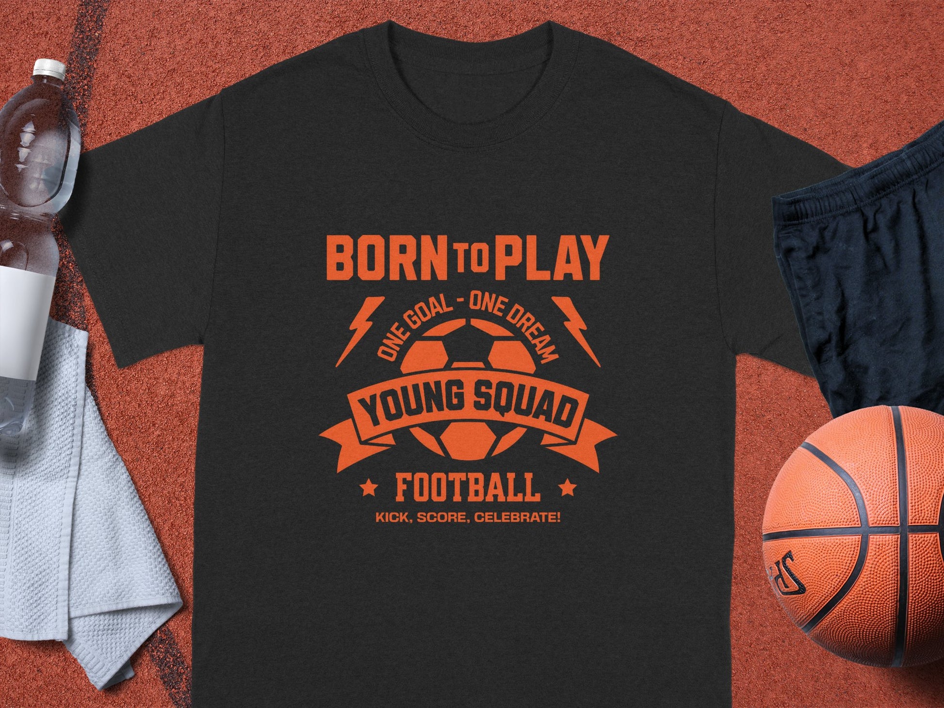 A black Garment Graphics football t-shirt with an orange Born to Play, One Goal - One Dream, Young Squad Football, Kick, Score, Celebrate! design is on a textured surface. Nearby are a basketball, towel, water bottle, and sports shoes—the ideal setup for passionate athletes.