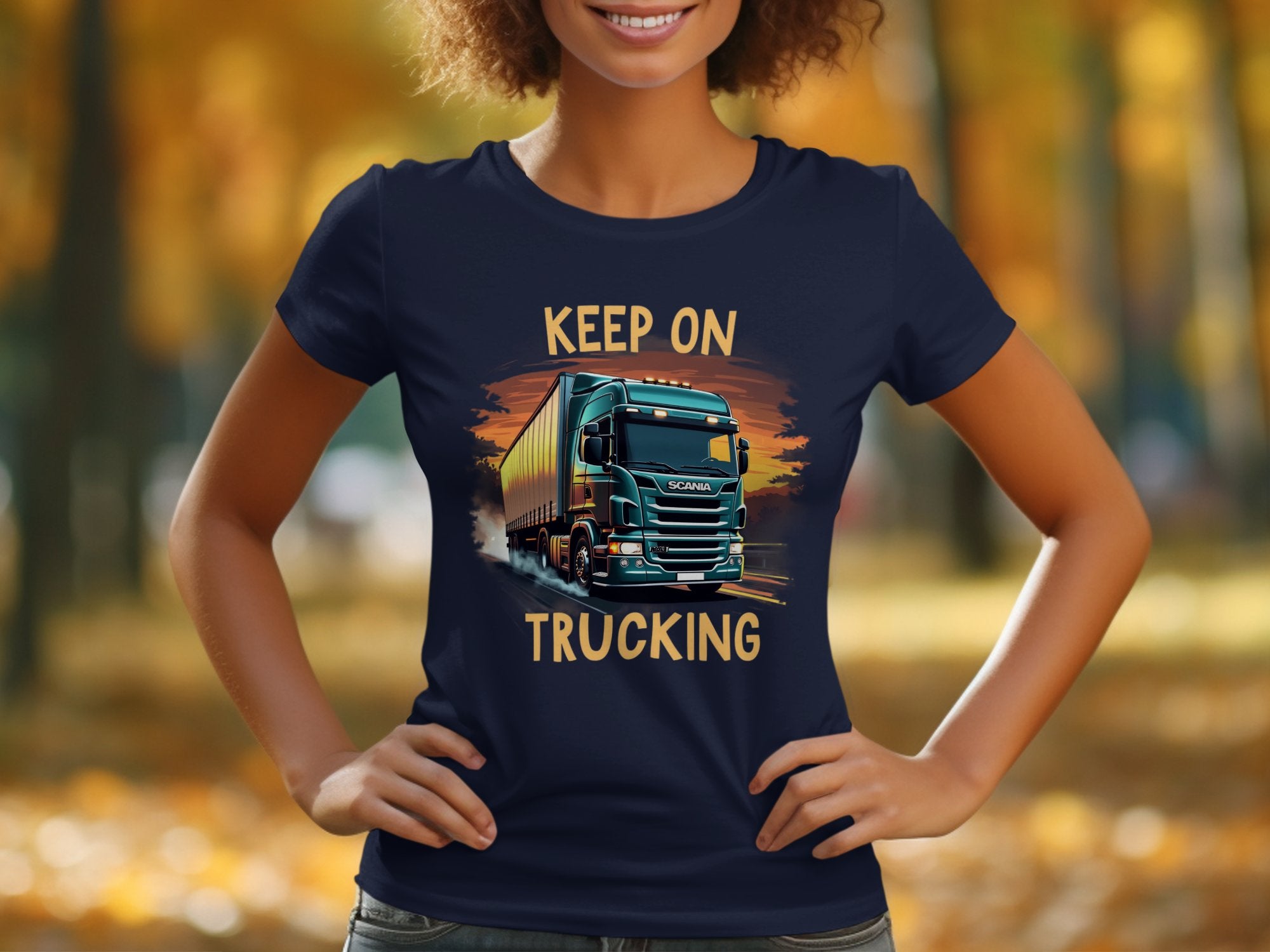 A person wearing a Garment Graphics navy T-shirt featuring a truck graphic and Keep On Trucking text, ideal for trucking enthusiasts, stands against blurred autumn foliage.