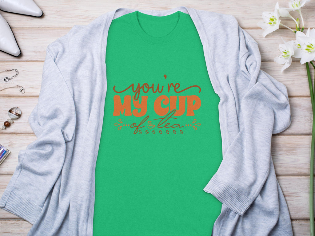 Garment Graphics Graphic T-shirt with Youre My Cup of Tea in red and black lettering on green 100% cotton fabric layers gracefully under a light gray cardigan. The look is enhanced with white flowers, glasses, and silver jewelry on a rustic wooden surface.