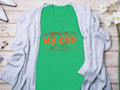 Garment Graphics Graphic T-shirt with Youre My Cup of Tea in red and black lettering on green 100% cotton fabric layers gracefully under a light gray cardigan. The look is enhanced with white flowers, glasses, and silver jewelry on a rustic wooden surface.