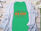 Garment Graphics Graphic T-shirt with Youre My Cup of Tea in red and black lettering on green 100% cotton fabric layers gracefully under a light gray cardigan. The look is enhanced with white flowers, glasses, and silver jewelry on a rustic wooden surface.