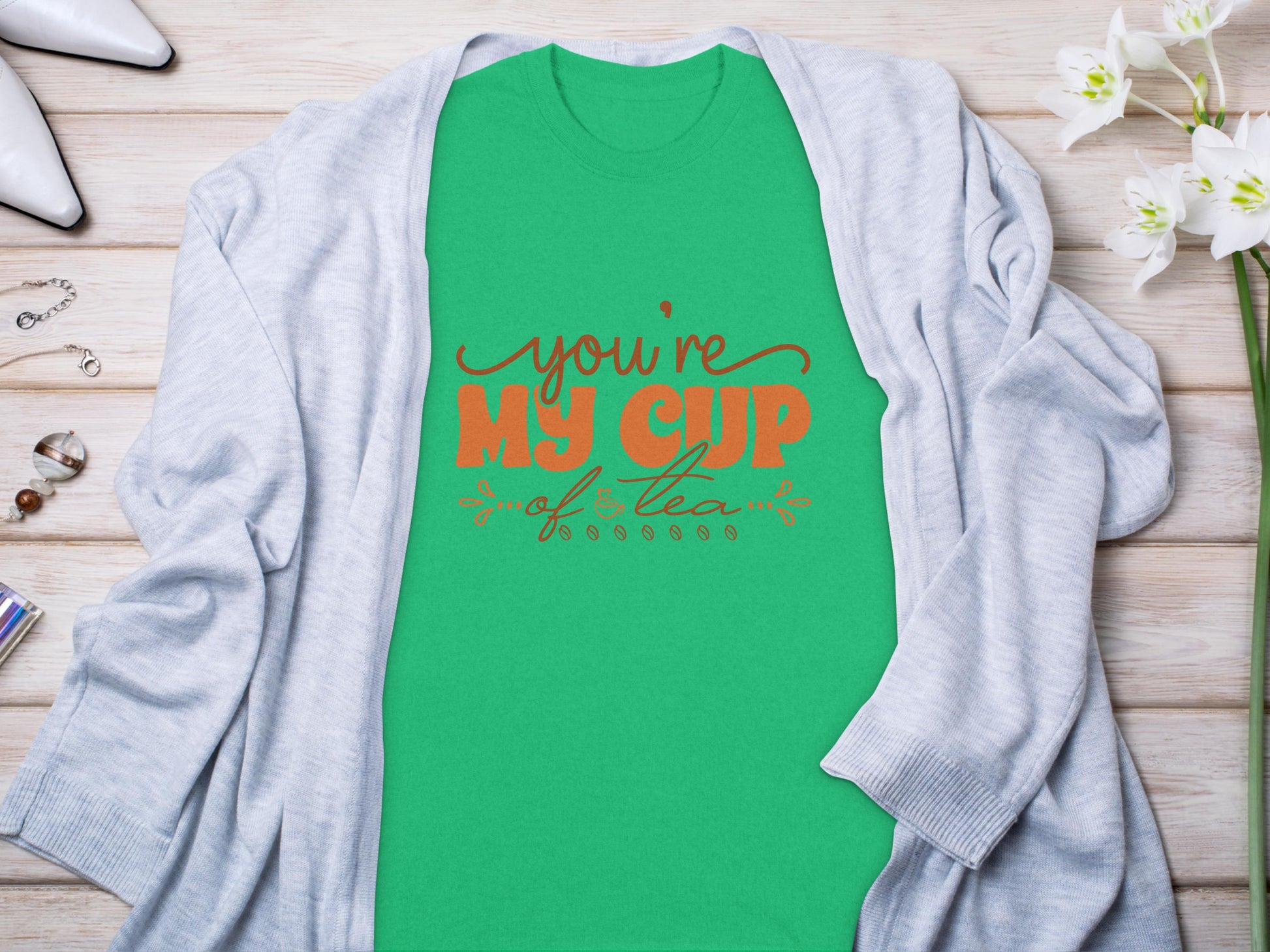 Garment Graphics Graphic T-shirt with Youre My Cup of Tea in red and black lettering on green 100% cotton fabric layers gracefully under a light gray cardigan. The look is enhanced with white flowers, glasses, and silver jewelry on a rustic wooden surface.