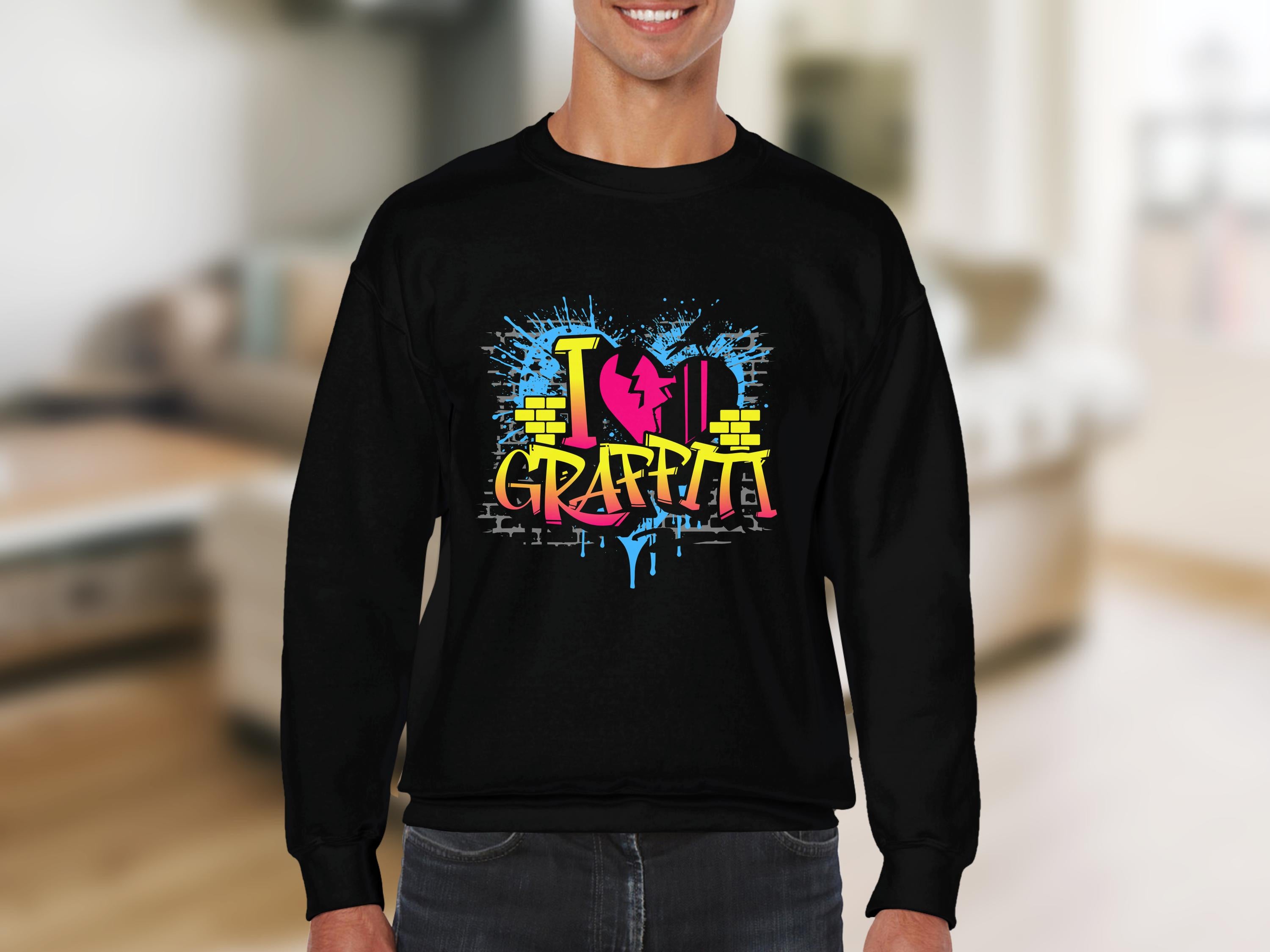 A person dons a Garment Graphics black sweatshirt featuring vibrant street art-style text saying I Love Graffiti, with a red heart and yellow brick design, set against a blurred indoor scene that emphasizes the lively and artistic vibe.