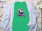 Garment Graphics classic fit green T-shirt features Soccer Mum in bold black and pink lettering with a soccer ball graphic. Displayed elegantly on wood, its paired with a gray cardigan, white heels, jewelry, and delicate white flowers for an eye-catching look.