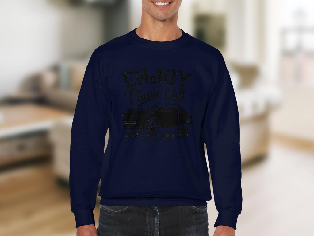 A person wears a dark blue ENJOY Classic Car sweatshirt by Garment Graphics, featuring a vintage automobile image. The softly blurred backdrop reveals a living room with beige furniture.