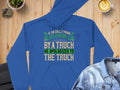 The Garment Graphics blue hoodie, featuring the witty quote If an Englishman gets run over by a truck, he apologizes to the truck in green, white, and yellow, is displayed on a wooden surface with a denim shirt, coffee cup, and potted plant.