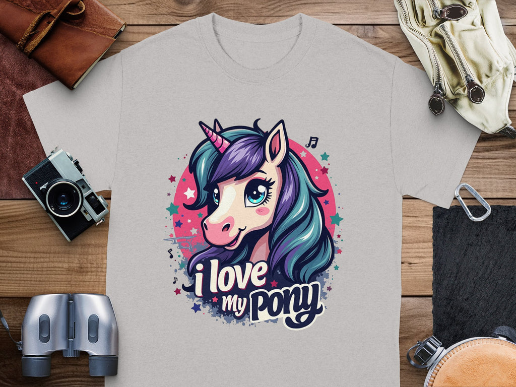 A gray Pony graphic T-shirt by Garment Graphics features a vibrant unicorn with purple and teal hair, displaying the phrase I love my pony. It’s perfect for unicorn enthusiasts and is pictured with a camera, binoculars, and leather pouch on a wooden surface.