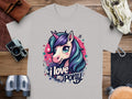 A gray Pony graphic T-shirt by Garment Graphics features a vibrant unicorn with purple and teal hair, displaying the phrase I love my pony. It’s perfect for unicorn enthusiasts and is pictured with a camera, binoculars, and leather pouch on a wooden surface.