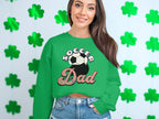 A person with long hair smiles, wearing a Garment Graphics green sweatshirt with a retro Soccer Dad design and soccer ball graphic. They stand in front of a wall decorated with green shamrocks, sporting denim shorts.