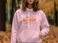 In a forest surrounded by cozy autumn moments, a person dons a light pink Garment Graphics hoodie with an enchanting youre my cup of tea design in orange. Blurred fall foliage and scattered leaves create the perfect backdrop.