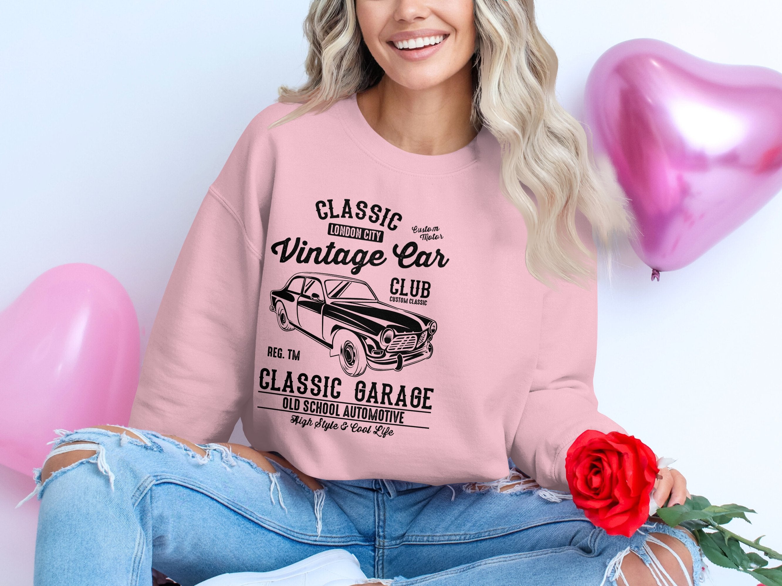 A woman wears a pink Garment Graphics sweatshirt with a vintage car graphic, exuding automotive charm. She smiles, holding a red rose amid pink balloons. Her ripped-knee jeans add to her casual yet chic look, reminiscent of classic London vibes.