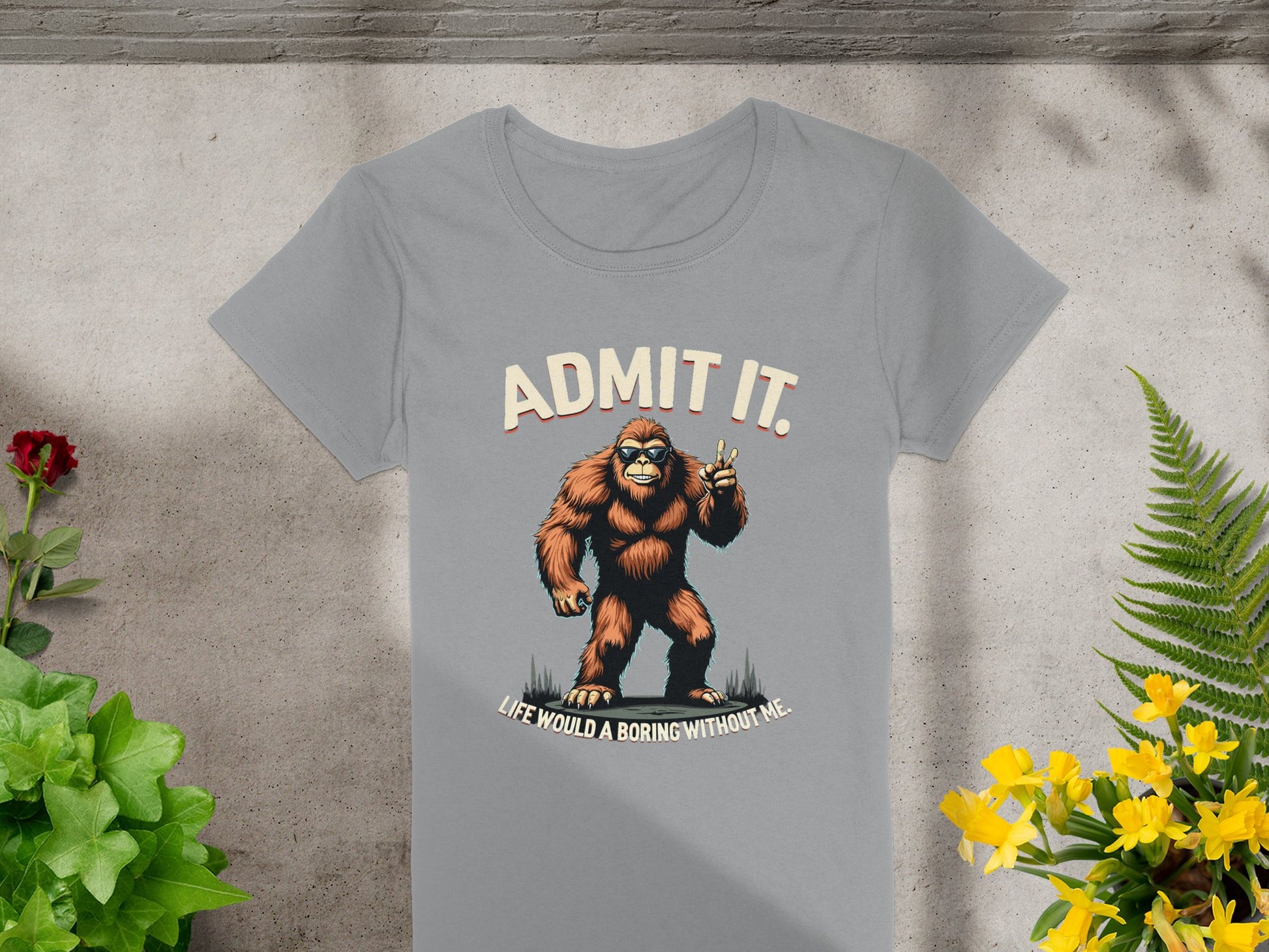 The Garment Graphics gray T-shirt features a cartoon Bigfoot with a coffee cup and the text Admit It. Life Would Be Boring Without Me. Its displayed flat on concrete, surrounded by green and yellow plants.