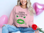 Clad in a Garment Graphics pink sweater with Happy St. Patricks Day and a green clover kiss design, a person with wavy blonde hair and ripped jeans sits holding a red rose, exuding festive spirit. Two pink heart-shaped balloons float nearby.