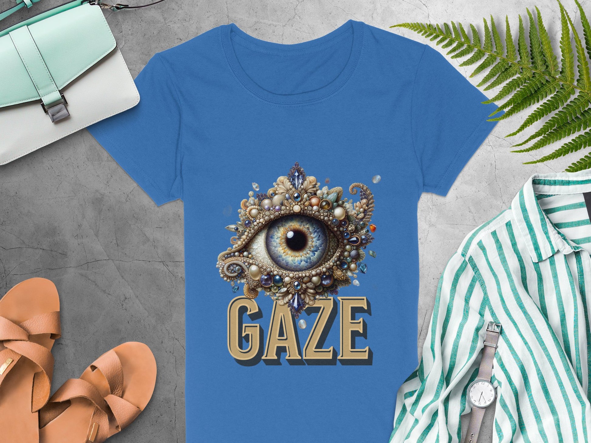 A blue Garment Graphics T-shirt featuring an eye design adds artistic flair to the scene, alongside a green-striped shirt, tan sandals, a white watch, a white and green clutch, and a fern—all elegantly arranged on a gray surface. Available in womens sizes.