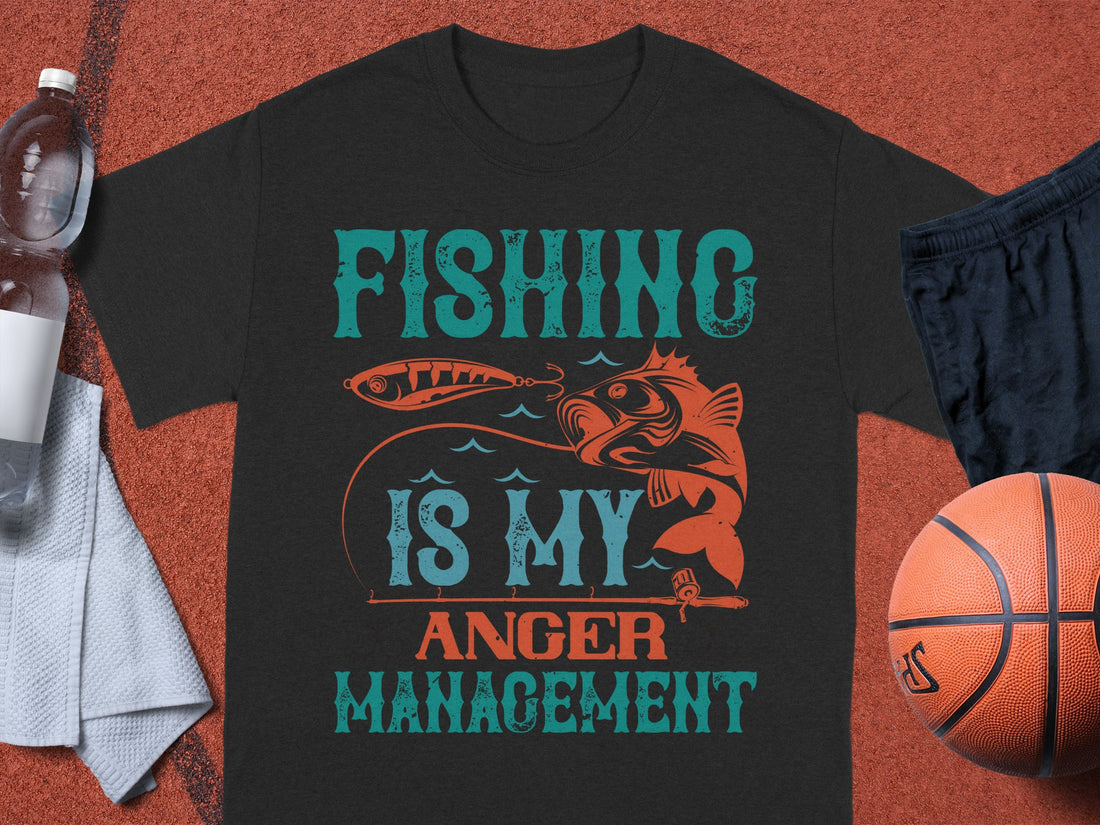 Garment Graphics black Fishing T-shirt for fishing lovers features Fishing Is My Anger Management in teal and orange, accented with a fish and lure design. It’s showcased on a basketball court paired with shorts, a basketball, towel, and water bottle.