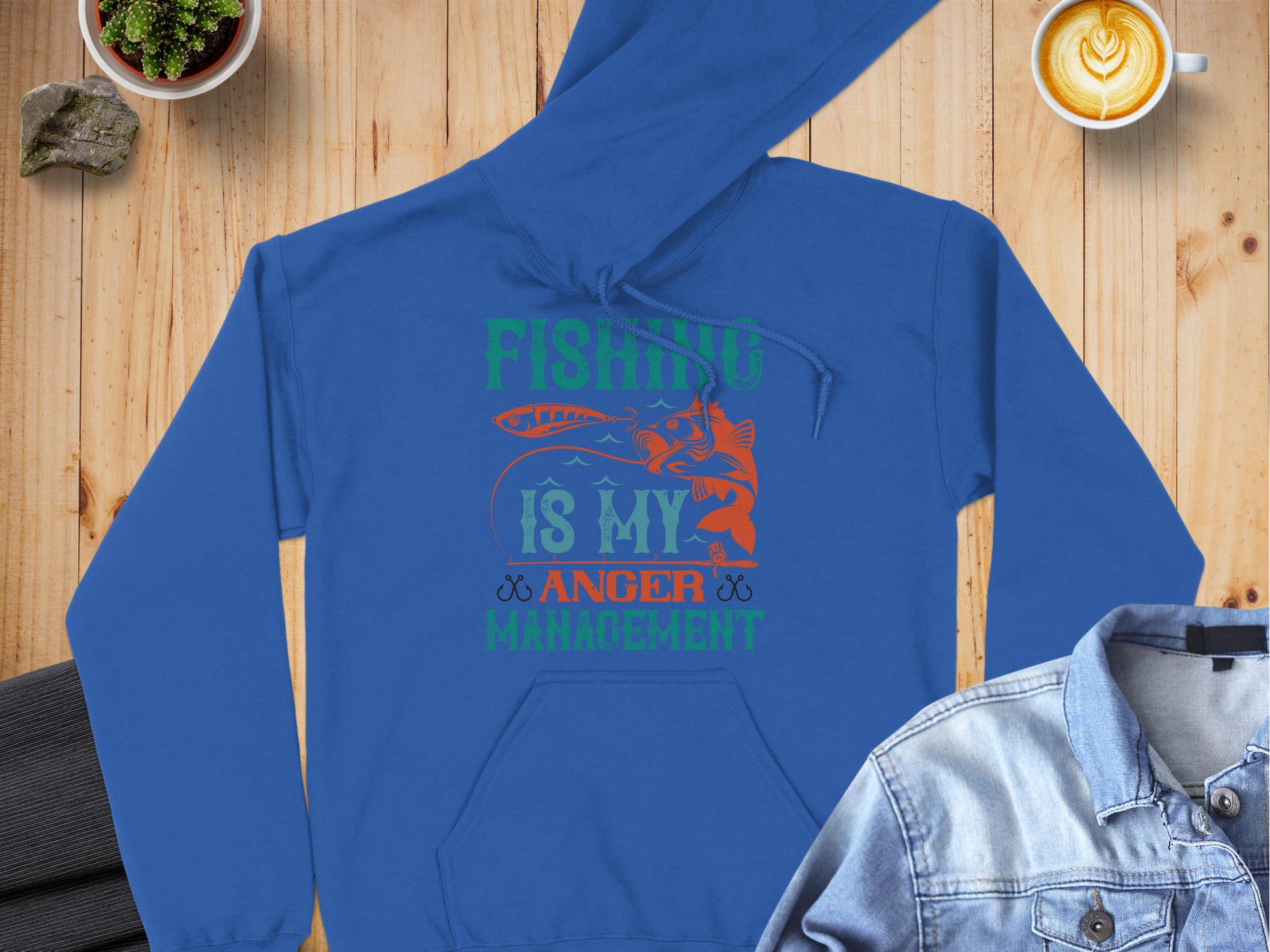 A blue hoodie from Garment Graphics Anglers Wardrobe displays Fishing is my anger management in green, orange, and white next to a fish graphic. It rests on wood surrounded by a jacket, coffee, and plant—ideal for those finding peace in fishing.