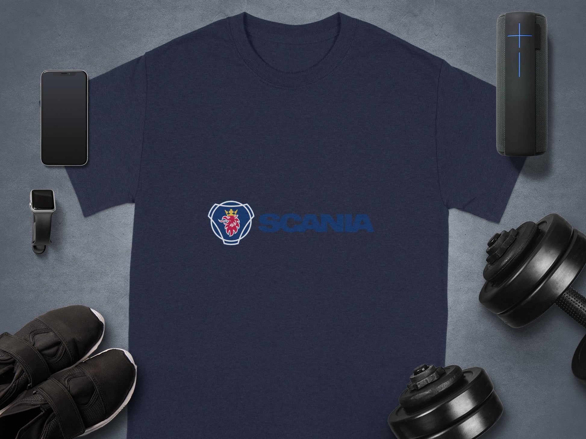 A navy blue cotton t-shirt by Garment Graphics features the Scania logo, flanked by a smartphone, smartwatch, portable speaker, dumbbells, and black sneakers on a gray surface—a must-have for Scania merchandise fans.