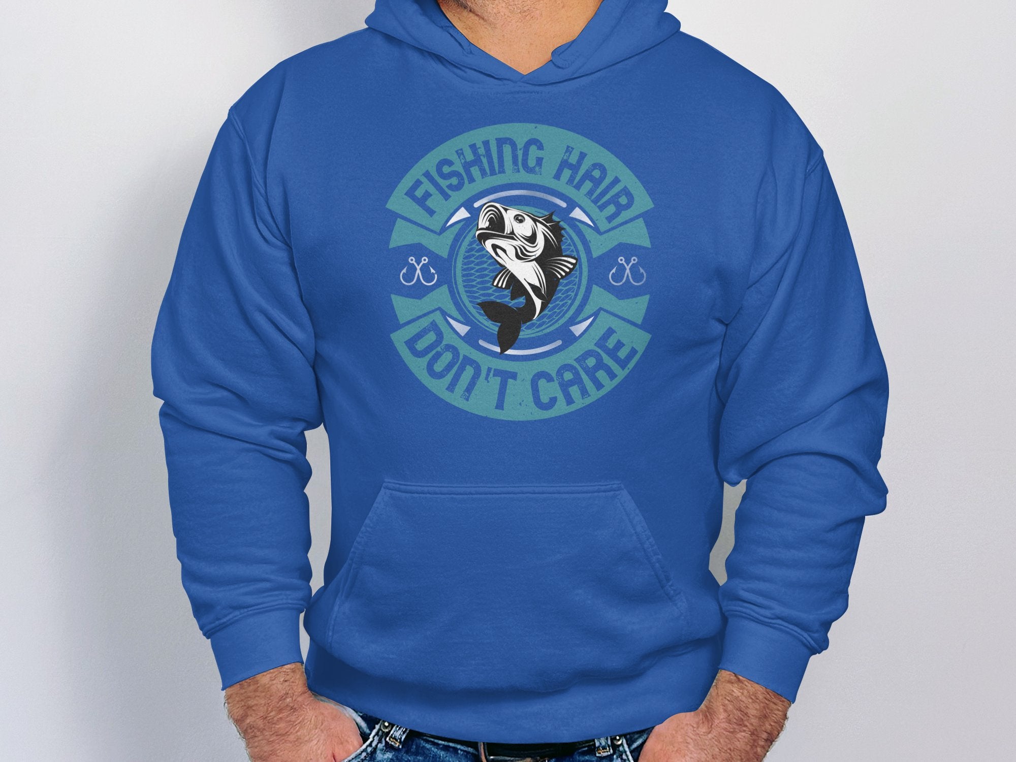 A person wears a Garment Graphics hoodie featuring the text Fishing Hair Dont Care around a fish image. Standing with hands in jeans pockets, they look poised for an angling adventure, with the focus on their torso.