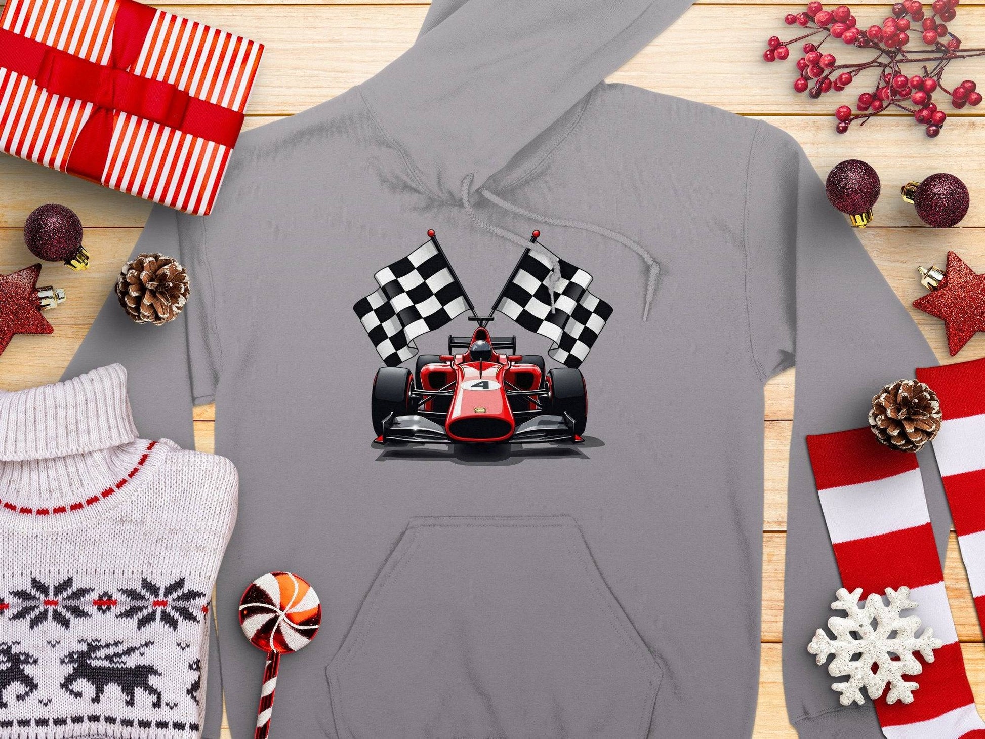 Garment Graphics presents a gray hoodie featuring a racing car with checkered flags, surrounded by holiday elements like wrapped gifts, pine cones, a Christmas sweater, candy, and red snowflakes set against a wooden background.