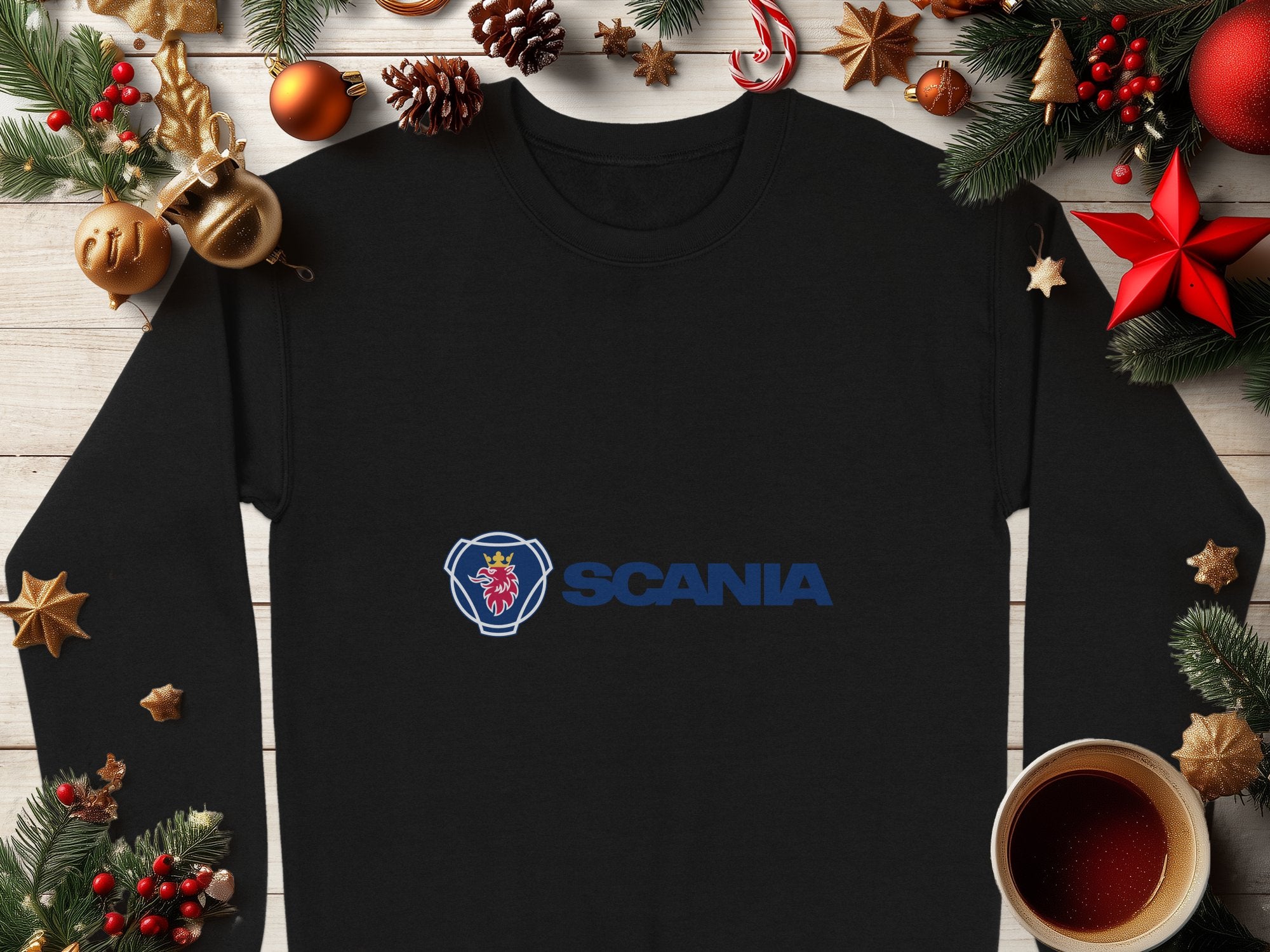 A black long-sleeve shirt from Garment Graphics with the Scania logo in blue and red lies on a wooden surface surrounded by holiday decor, including pine branches, pine cones, red stars, and hot chocolate. Ideal for casual outings, its unisex sizing offers a comfortable fit for everyone.
