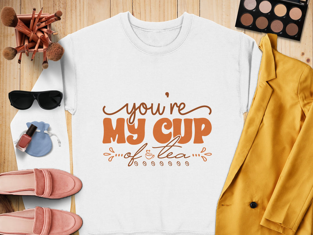 Flat lay features Garment Graphics white shirt with Youre My Cup of Tea in orange, paired with a yellow blazer, pink shoes, sunglasses, makeup, and a blue pouch. This charming ensemble looks cozy yet chic on the wooden surface.