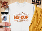 Flat lay features Garment Graphics white shirt with Youre My Cup of Tea in orange, paired with a yellow blazer, pink shoes, sunglasses, makeup, and a blue pouch. This charming ensemble looks cozy yet chic on the wooden surface.