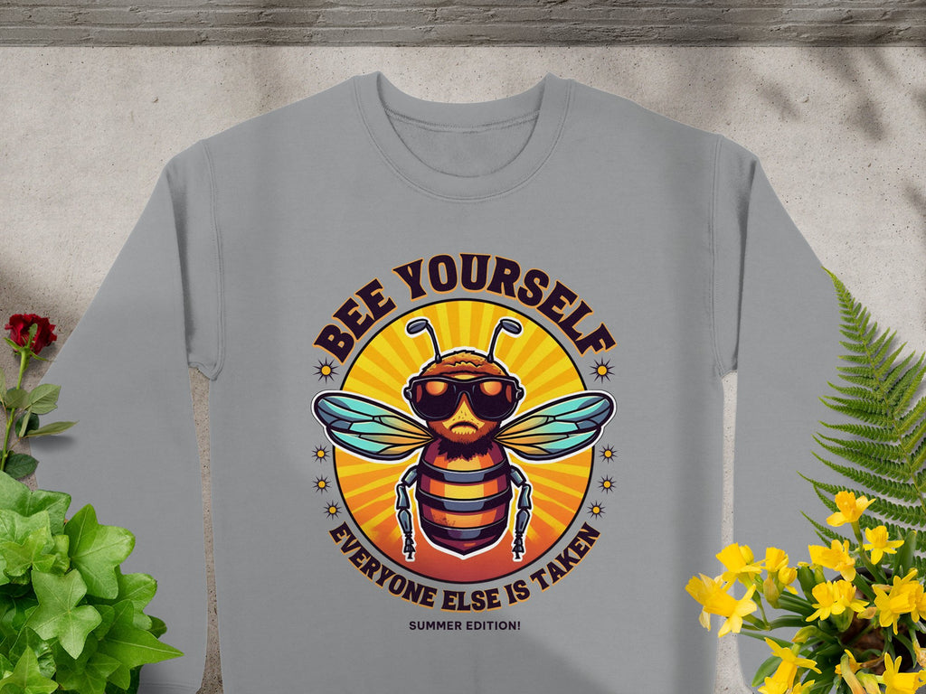 Garment Graphics gray sweatshirt showcases a bee in sunglasses giving a thumbs up, with the text Bee Yourself above Everyone Else Is Taken. Part of the Summer Edition, this trendy unisex piece seamlessly complements any wardrobe, surrounded by lush plant designs.