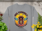 Garment Graphics gray sweatshirt showcases a bee in sunglasses giving a thumbs up, with the text Bee Yourself above Everyone Else Is Taken. Part of the Summer Edition, this trendy unisex piece seamlessly complements any wardrobe, surrounded by lush plant designs.