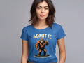 A brown-haired woman wears a Garment Graphics blue T-shirt featuring the playful Admit It Bigfoot design, with a Bigfoot illustration and the text, ADMIT IT. Life would be boring without me, all set against a plain gray backdrop.